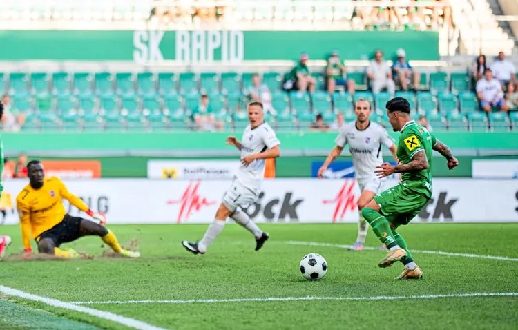 Ein Fußballspieler im grünen Trikot des SK Rapid kurz vor einem Torschuss während eines Spiels. Der Torwart in gelbem Trikot versucht, den Schuss zu blockieren, während Spieler der gegnerischen Mannschaft im weißen Trikot zuschauen. Zuschauer sind im Hintergrund auf den Tribünen sichtbar.
